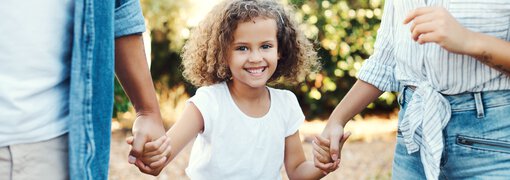 Ein kleines Mädchen mit braunen lockigen Haaren wird von seinen Eltern an beiden Händen gehalten | © PeopleImages - Getty Images/iStockphoto